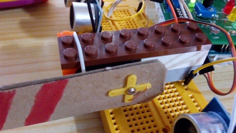 Detalle de la unión barrera hecha con cartón y el servo, sujetado con piezas de lego y la barrera utilizando el accesorio cruz del servo y atado con un hilo de cobre